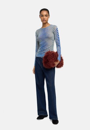 Kobieta ubrana w niebieską, kropkowaną bluzkę z długim rękawem i ciemne dżinsy, trzymająca dużą, puszystą czerwoną torebkę kopertówkę, stojąca na jednolitym tle.