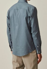 Chemise homme à manches longues en tissu bleu doux, dotée d'un col classique, de poignets boutonnés et d'un ourlet arrondi. Texture lisse, sans motifs.