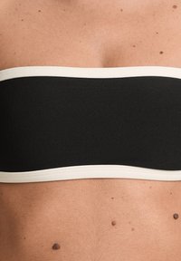 Črn bandeau top s širokim kremnim obrobom, izdelan iz gladkega materiala. Ima preprost dizajn brez dodatnih vzorcev ali okrasnih elementov.