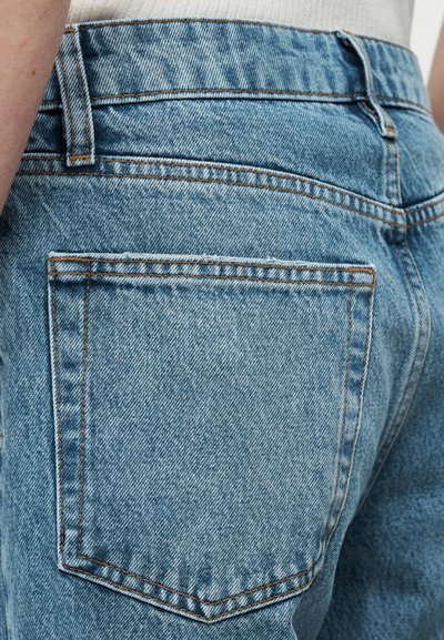 Κοντινή λήψη της πίσω τσέπης από ανοιχτό μπλε τζιν με εμφανείς ραφές και θηλιά ζώνης, φορεμένη από άτομο που φοράει λευκό ριμπ μπλουζάκι.