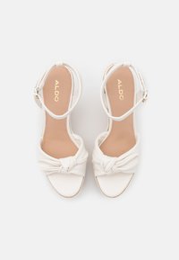 Witte sandaal met hak, voorzien van een geknoopt design op de teen, een textuurvormige binnenzool en een smalle enkelband met een gesp sluiting.