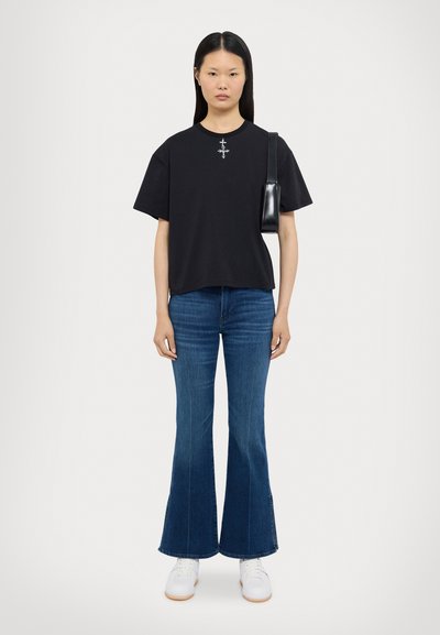 Sort kortærmet t-shirt med et centralt grafisk motiv, parret med flagrende blå jeans og hvide sneakers. Modellen holder en lille sort håndtaske.