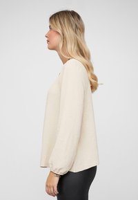 Beige lang mouw blouse met een gestructureerde stof, losse pasvorm en elastische manchetten. De halslijn heeft een subtiele opening. Draag het over zwarte leggings.