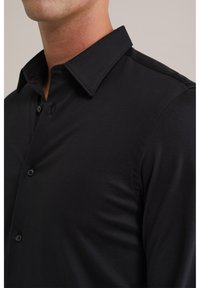 Juoda marškinėliai su mygtukais, turintys struktūruotą apykaklę, lygią audinio tekstūrą ir subtilias siuvimo detales. Artimas marškinėlių ir apykaklės vaizdas.