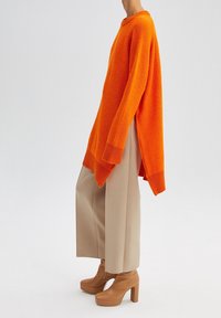 Przewymiarowany pomarańczowy sweter z prążkowanego materiału, z długimi rękawami i bocznymi rozcięciami, połączony z beżowymi szerokimi spodniami i brązowymi botkami na platformie.