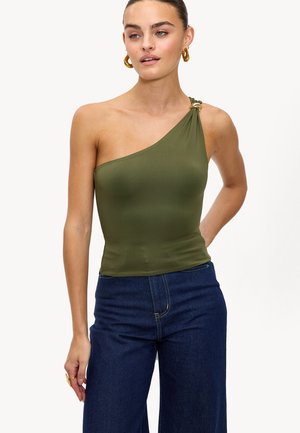 Femme portant un haut une épaule vert olive avec un détail en anneau doré et un jean taille haute bleu foncé, se tenant avec une main derrière le dos.