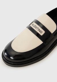 Melna un krēmkrāsas lakotas ādas kurpe ar noapaļotu purngali, kurai ir plata melna siksna un sudraba "Valentino" etiķete.
