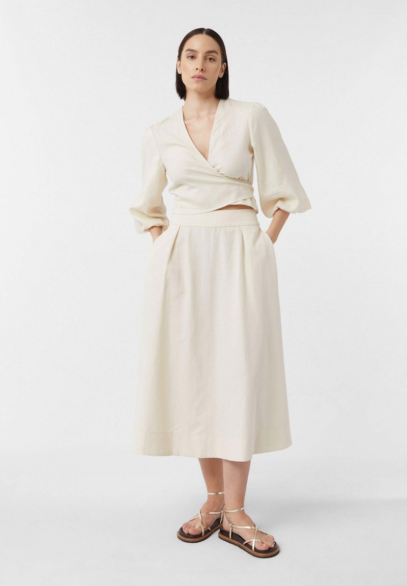 Cremefarbenes Wickeltop mit langen Ärmeln, kombiniert mit einem passenden bodenlangen Rock und Sandalen mit goldenen Riemen. Weicher, strukturierten Stoff.