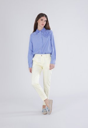 Žena stojící, oblečená v modrobíle pruhované košili na knoflíky s mašlí, krémové kalhoty a béžové mokasíny s modrými tkaničkami.