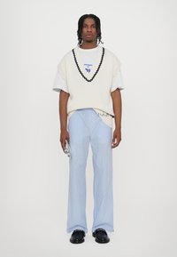 Gilet bianco a maglia con bordi neri sopra una maglietta bianca, abbinato a pantaloni a gamba larga a righe blu chiaro e scarpe nere.