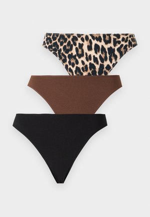 Femme debout pieds nus portant un soutien-gorge noir et un sous-vêtement style bikini à imprimé léopard sur un fond blanc uni.