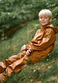 Bruin waterdicht regenpak gemaakt van glanzend materiaal, voorzien van een capuchon en elastische manchetten, met een logo-accent op de mouw. Kind zittend buiten.