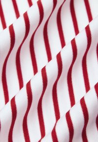 Biała tkanina z równomiernie rozmieszczonymi pionowymi czerwonymi paskami, pokazująca delikatne fałdy i szczegóły tekstury.