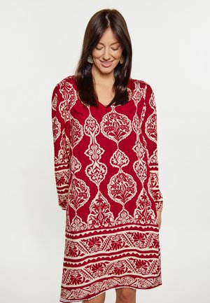 Femeie cu păr închis la culoare care poartă o rochie roșie cu modele florale și geometrice albe, uitându-se în jos și zâmbind pe un fundal simplu.