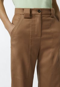 Smeđe krojene hlače od glatkog materijala, širokog kroja, sa prednjim džepovima i zatvaranjem na dugme uz pojas s petljama.