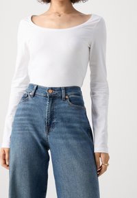 Kvinna som bär en vit långärmad topp med rundad hals och inknäppt i högmidjade blå denimjeans mot en enkel bakgrund.