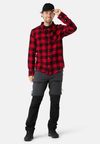 Rood en zwart geblokt flanellen shirt, grijze cargobroek met ritsen, zwarte pet en zwarte wandelschoenen. Casual outdoor kleding.
