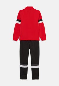 Trenerka z rdečo jakno z ovratnikom in črnimi ter belimi črtami na rokavih, skupaj s črnimi hlačami s svetlečimi črtami ob gležnjih.