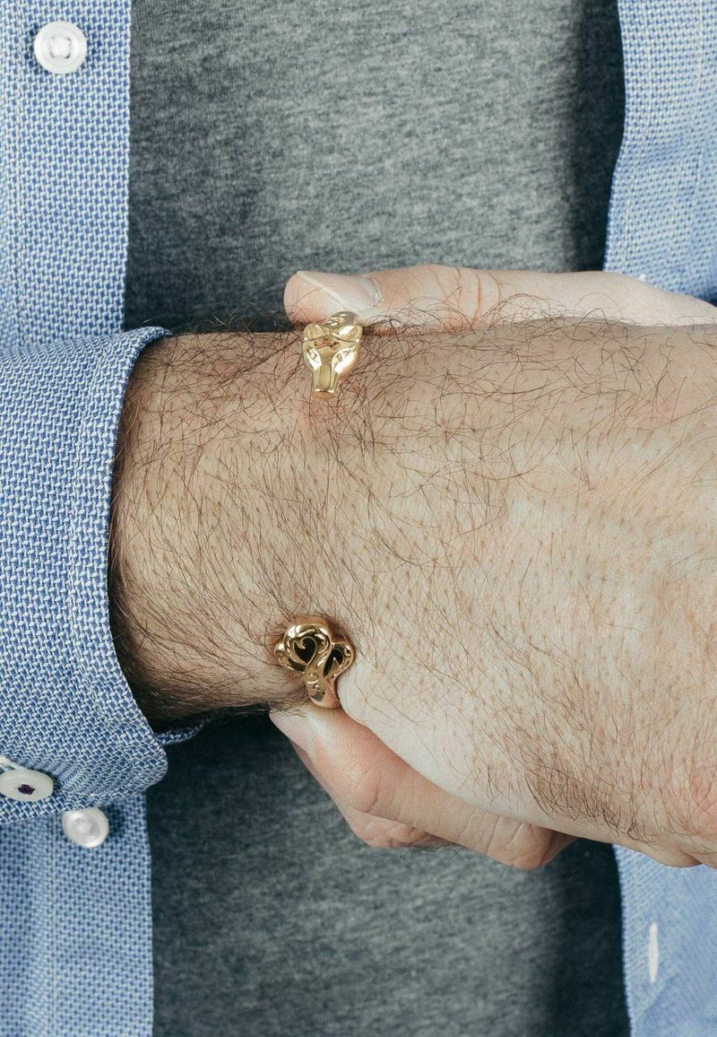 Pulseras en forma de animales en tono dorado con un detallado intrincado, lucidas en una muñeca, contrastando con una camisa gris y el puño de una camisa a cuadros azul.