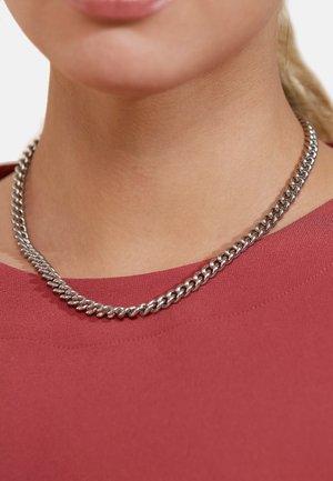 Silver metal chain necklace with twisted links, resting on a neck that is partially visible. Background features a red garment.