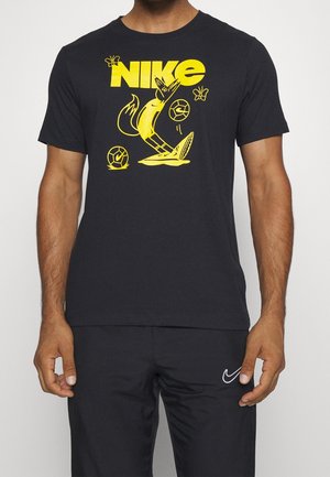 Mann trägt ein schwarzes Nike-T-Shirt mit einem gelben Fuchs, der Fußball spielt, zwei Fußbällen und Schmetterlingen, dazu schwarze Nike-Hosen.