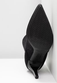 Zapato de tacón alto negro con parte superior de tela texturizada y puntera afilada. La suela presenta un diseño patrón para mayor agarre.