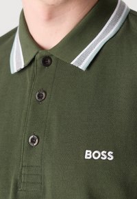 Gros plan d'un polo vert avec trois boutons, col rayé blanc et logo blanc "BOSS" brodé sur la poitrine.