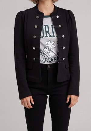 Femme portant une veste militaire noire avec des boutons argentés par-dessus un t-shirt gris à motif de palmiers et texte.