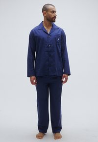 Σετ πυτζάμες σε σκούρο μπλε χρώμα, κατασκευασμένο από απαλό ύφασμα, με πουκάμισο με κουμπιά και μακριά μανίκια και ταιριαστό παντελόνι, με λεπτομέρεια τσέπης.