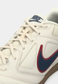 Buty Nike z beżową, syntetyczną cholewką z granatowymi akcentami, charakterystycznym czerwonym logo oraz kremowymi sznurowadłami. Podeszwa wykonana z gumy z teksturowanym wykończeniem.