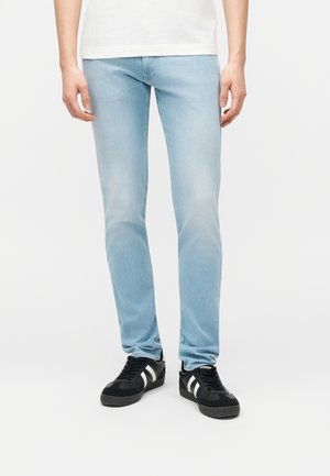 Personne portant un jean slim bleu clair et des baskets noires à rayures blanches, debout devant un fond blanc uni.