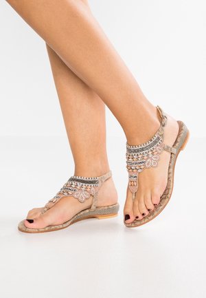 Pieds portant des sandales plates beiges ornées de perles et de bijoux complexes, avec un vernis foncé sur les orteils, sur fond blanc.