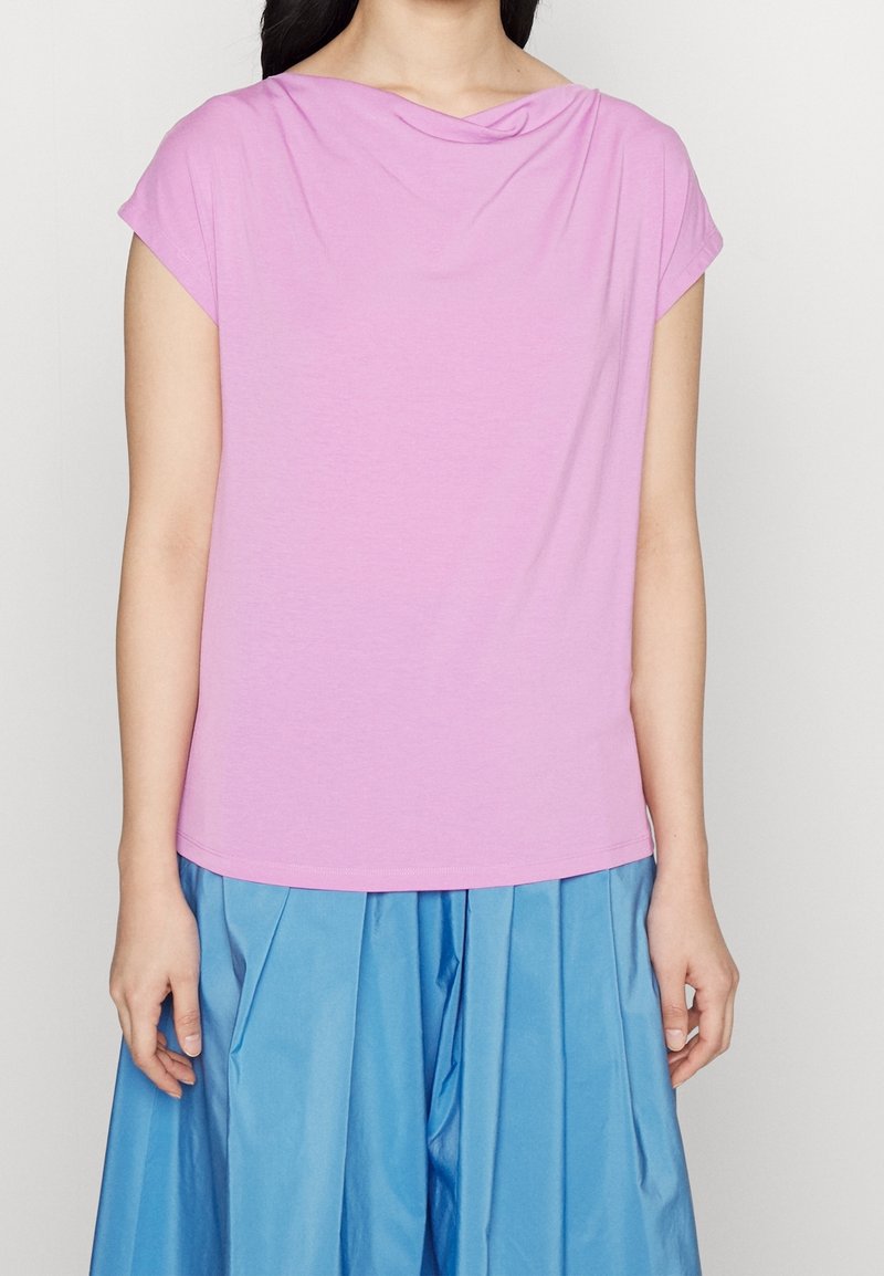 Femme portant un haut à manches courtes violet clair avec un col drapé et une jupe plissée bleue ample, debout devant un fond uni.