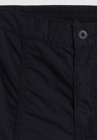 Pantalon noir à taille élastique avec fermeture par bouton et coutures visibles sur le devant, montrant une partie de la poche et de la braguette.