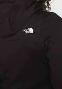 Personne portant un sweat à capuche noir North Face avec le logo de la marque imprimé en blanc sur le haut du dos, près de l'épaule.