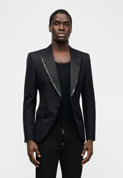 Άνδρας που φοράει μαύρο σακάκι του σμόκιν με σατέν πέτο πάνω από μαύρο πουκάμισο και μαύρο παντελόνι, στέκεται μπροστά σε απλό ανοιχτό φόντο.