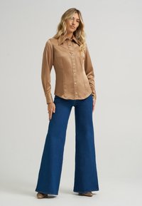 Μακρυμάνικη, πουκαμίσα με κουμπιά από σατέν ύφασμα, καμηλό χρώμα με pointed collar και καμπυλωτό στρίφωμα; συνδυασμένη με μπλε φλαρέ παντελόνια και χρυσά παπούτσια.