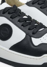 Černobílé kožené tenisky s kulatým logem, texturovanými černými tkaničkami a perforacemi na špičce pro prodyšnost.