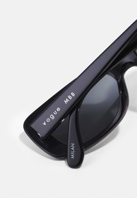 Óculos de sol pretos com acabamento brilhante, design angular, lentes escuras e inscrições brancas "vogue MBB" e "MILAN" nas hastes.