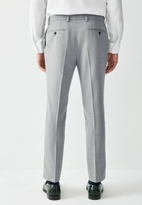 Grijze broek van een gestructureerde stof, met een getailleerde pasvorm, een middenvouw en twee achterzakken. Gecombineerd met glanzende zwarte schoenen.