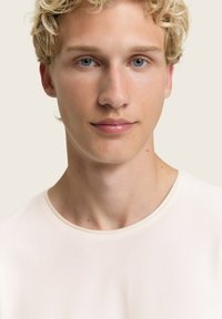 Junge Person mit kurzem, lockigem, blonden Haar, blauen Augen, die ein einfaches weißes Rundhalsoberteil trägt und mit neutralem Ausdruck nach vorne blickt.