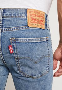 Un primer plano del bolsillo trasero de unos vaqueros Levi's azules con la etiqueta de marca roja y la etiqueta de cuero de Levi Strauss & Co., usados por una persona con una camisa blanca.