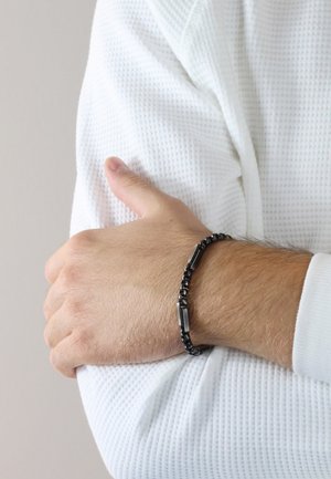 Black metal bracelet with interlocking links and rectangular accents, worn on a wrist, against a textured white textile background.