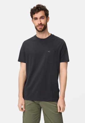 Mand med skæg iført mørkegrå kortærmet T-shirt med lomme og olivengrønne bukser, stående mod en ensfarvet lys baggrund.