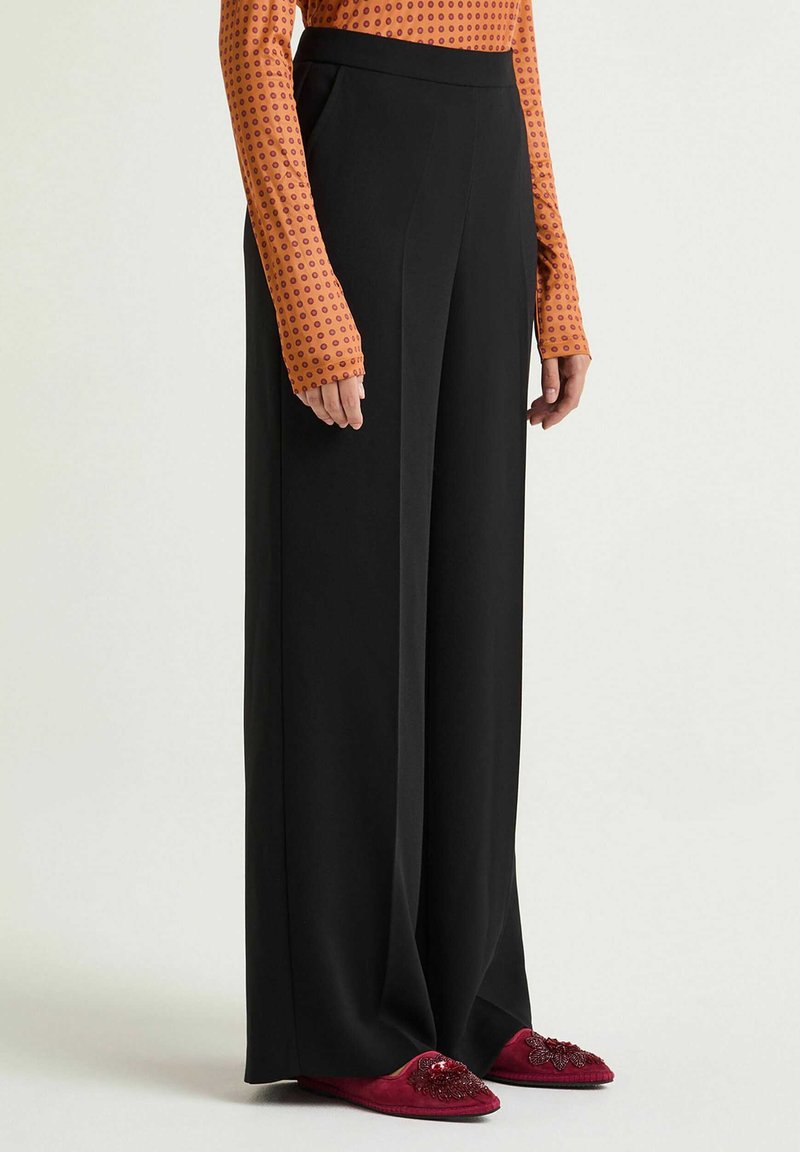 Pantalon noir à jambes larges en tissu lisse avec une taille haute. Associé à un haut à manches longues à motifs orange et rouge, chaussures ornées.