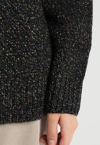 Dark, textured knit sweater featuring multicolored thread accents, ribbed cuffs, and a slightly loose fit, contrasting with a light beige background.