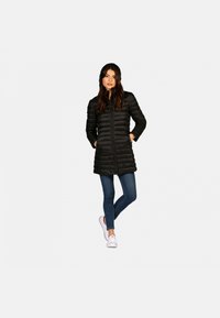 Doudoune noire avec fermeture éclair, dotée d'une capuche, de manches longues et de coutures horizontales. Associée à un jean bleu et des baskets blanches.