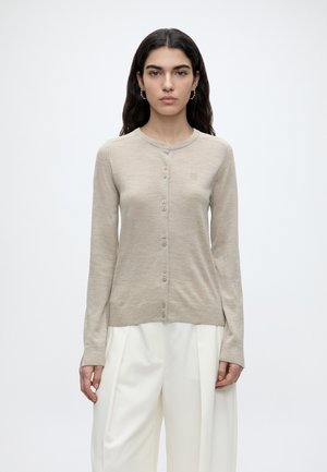 Femme aux longs cheveux foncés portant un cardigan beige à boutons manches longues et un pantalon blanc à jambes larges, debout devant un fond clair uni.