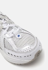 Fehér sportcipő hálós felsőrésszel, struktúrált mintákkal, ezüst részletekkel és szürke fűzőkkel. Kiemelkedő N logó a nyelvén.