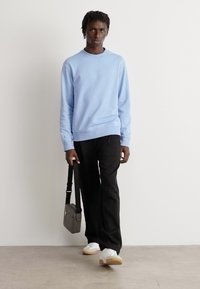 Sudadera de crewneck azul claro, pantalones negros, zapatillas blancas. El modelo lleva un bolso mensajero gris. Diseño simple, corte relajado.